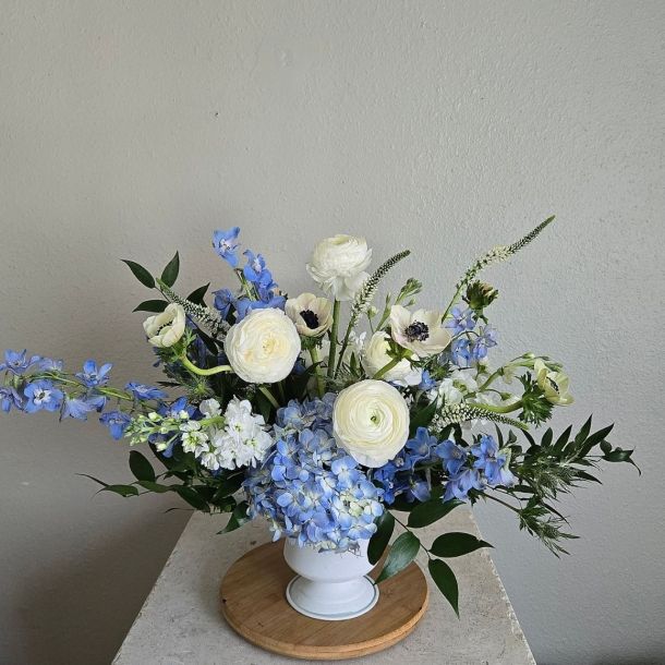 Naomi floral arrangement with blue hydrangeas, delphinium, and white ranunculus in a white pedestal vase